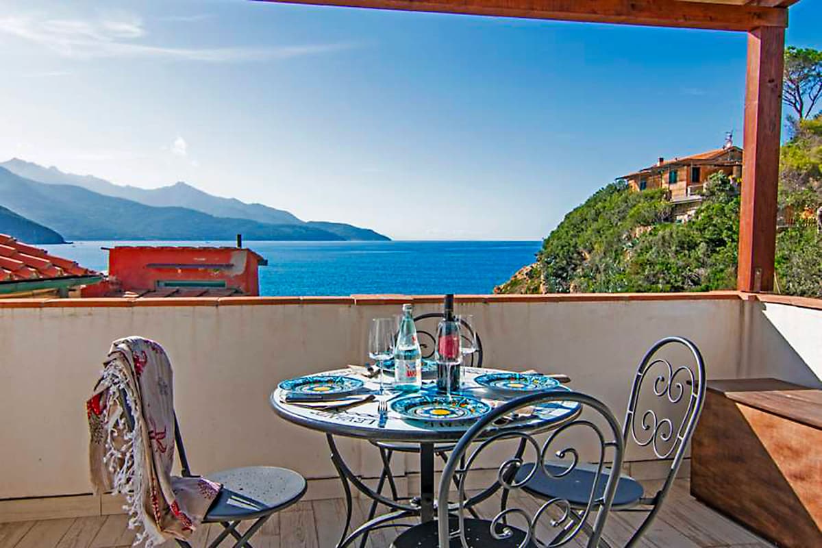 A terrace with a scenic ocean view features a round table set for dining. Four ornate metal chairs surround the table, complemented by glasses and plates. The backdrop showcases distant mountains and a clear blue sky, creating a serene outdoor setting.