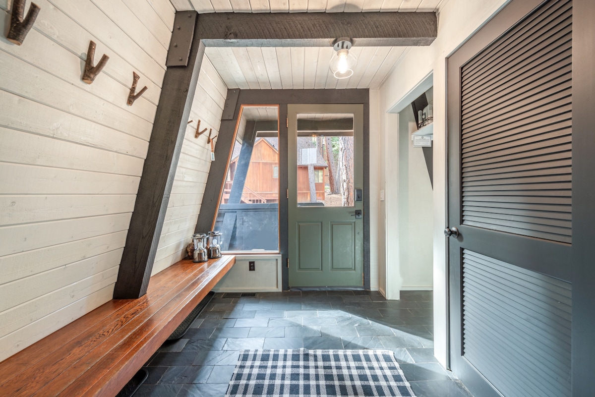 An entryway is displayed with a slate floor and whitewashed wooden walls. A long wooden bench runs along one side, with hooks for hanging items above. Natural light enters through a large front door, creating a welcoming atmosphere.