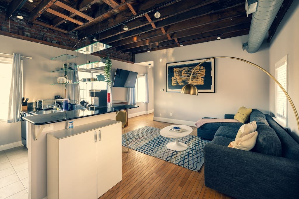 A spacious living area features exposed wooden beams and an open kitchen design. A dark couch is complemented by colorful pillows. A round coffee table rests on a patterned rug, while sunlight streams through two windows, highlighting the warm wooden flooring.