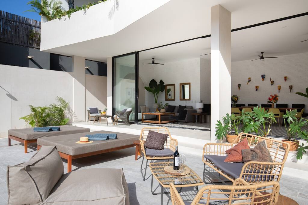 An outdoor seating area is presented with comfortable lounge chairs and a small table, surrounded by lush greenery. Large glass doors illuminate the interior space, showcasing a stylish living area with a dining table in the background.