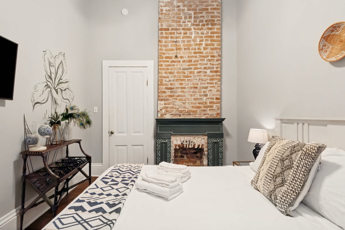 A bright sleeping area highlights exposed brick accents and a decorative fireplace. A spacious bed is adorned with plush pillows and neatly arranged towels. Natural light fills the room, while a small side table and plant add subtle touches of comfort.