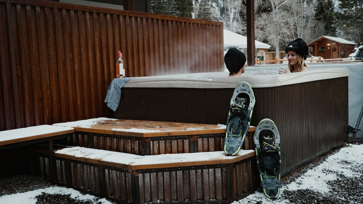 A heated outdoor hot tub is framed by wooden decking, featuring steps leading up to it. Snow-covered ground surrounds the area, and snowshoes are placed casually at the base of the steps, adding to the winter ambience.
