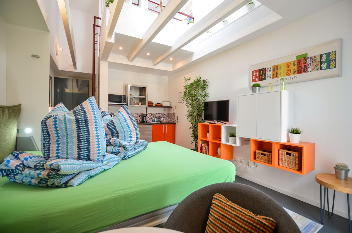 A spacious sleeping area features a large, green bed covered with a textured blue and white blanket. Above, skylights allow natural light to fill the room, while a kitchenette with modern appliances is visible in the background. Decorative plants and colorful shelves add a touch of warmth.