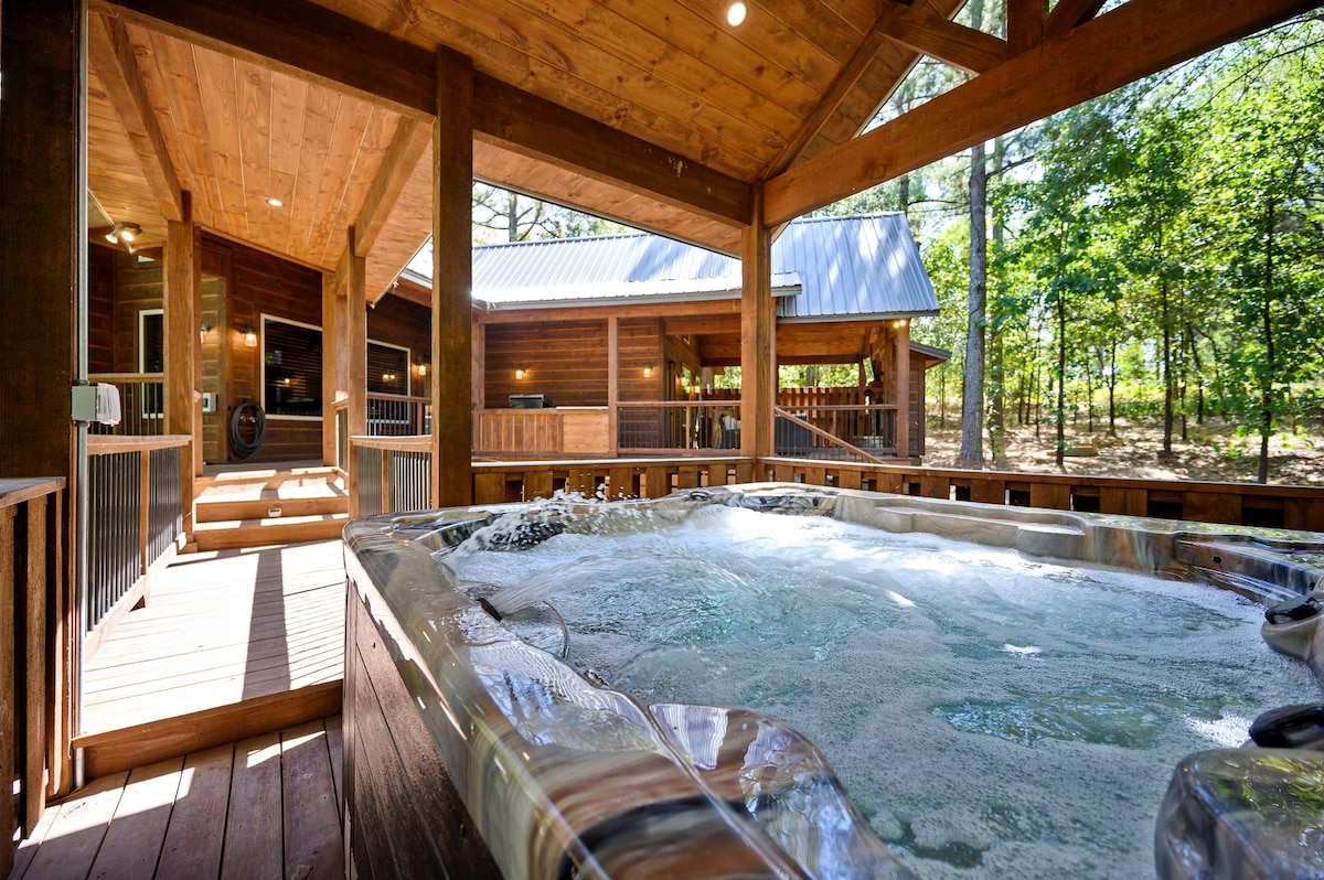 A spacious outdoor area features a large hot tub beneath a wooden-covered structure, surrounded by trees. The deck has natural wood finishes and provides access to the cabin and a serene, private atmosphere.