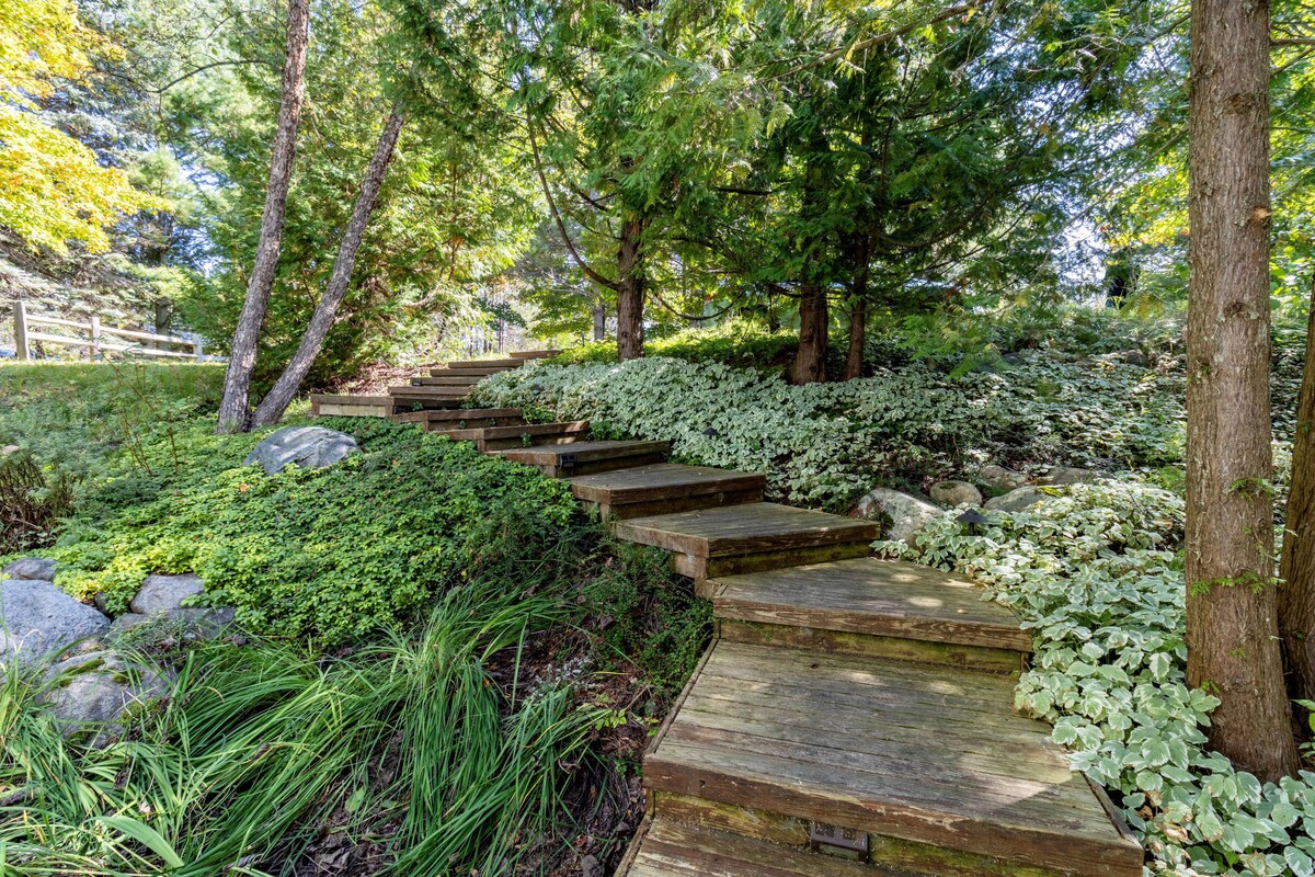 Wooden steps are surrounded by lush greenery, leading through a canopy of trees. The staircase is framed by shrubs and stones, inviting exploration and connection with nature. Sunlight filters through the leaves, creating a serene outdoor pathway.