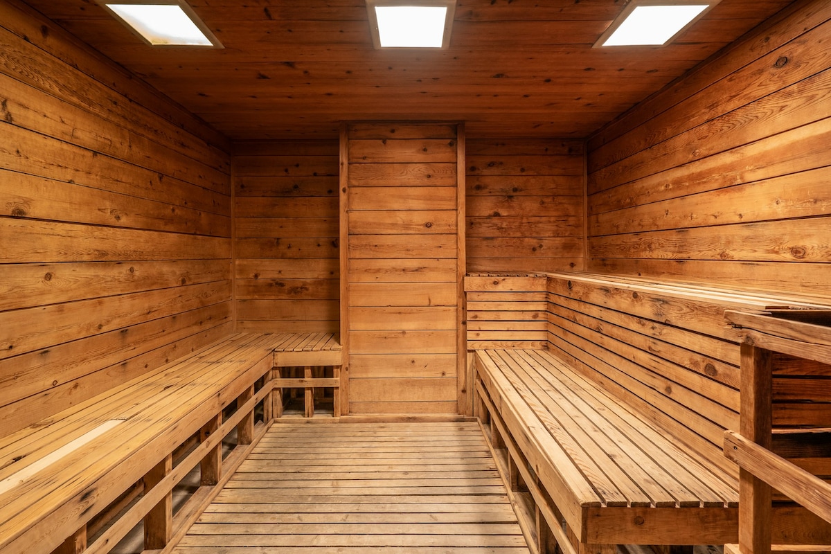 A wooden sauna interior is displayed, featuring a slatted bench along two sides and a central space. The walls are constructed from warm wood panels, and the ceiling is illuminated by four square lights, enhancing the serene environment.