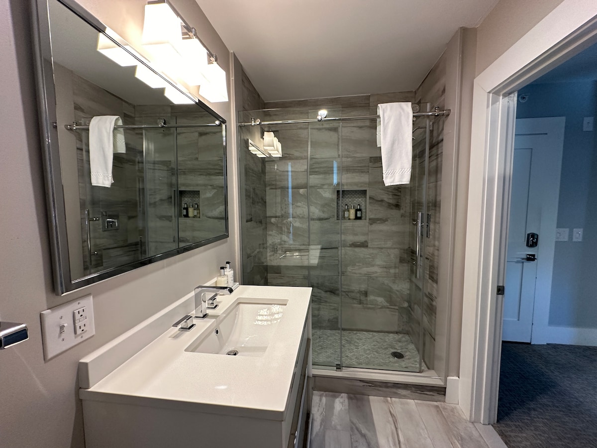 A modern ensuite bathroom features a spacious walk-in glass shower and a white vanity with dual sinks. A large mirror reflects the sleek design, and clean towels hang neatly on display. Neutral tones and a contemporary aesthetic create an inviting space.