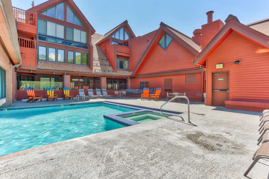 The outdoor area features a heated pool bordered by lounge chairs. A hot tub is adjacent to the pool. The surrounding buildings display a rustic red exterior and large windows, offering ample natural light.