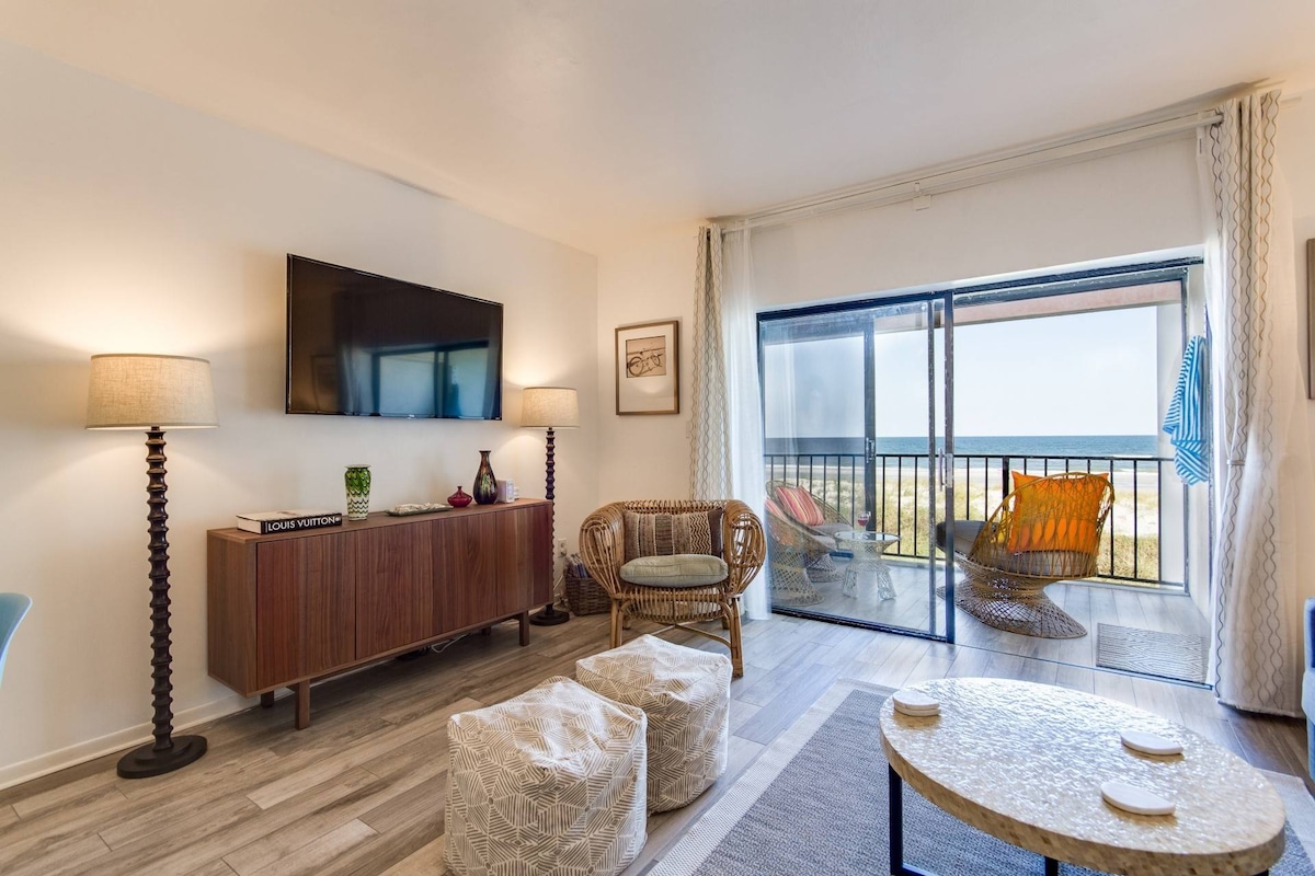 A well-furnished living area features natural light streaming through sliding glass doors, offering views of the beach. A spacious media console holds a flat-screen TV, while cozy seating options include a patterned chair and woven lounge chairs. Soft poufs and a textured coffee table enhance the inviting atmosphere.