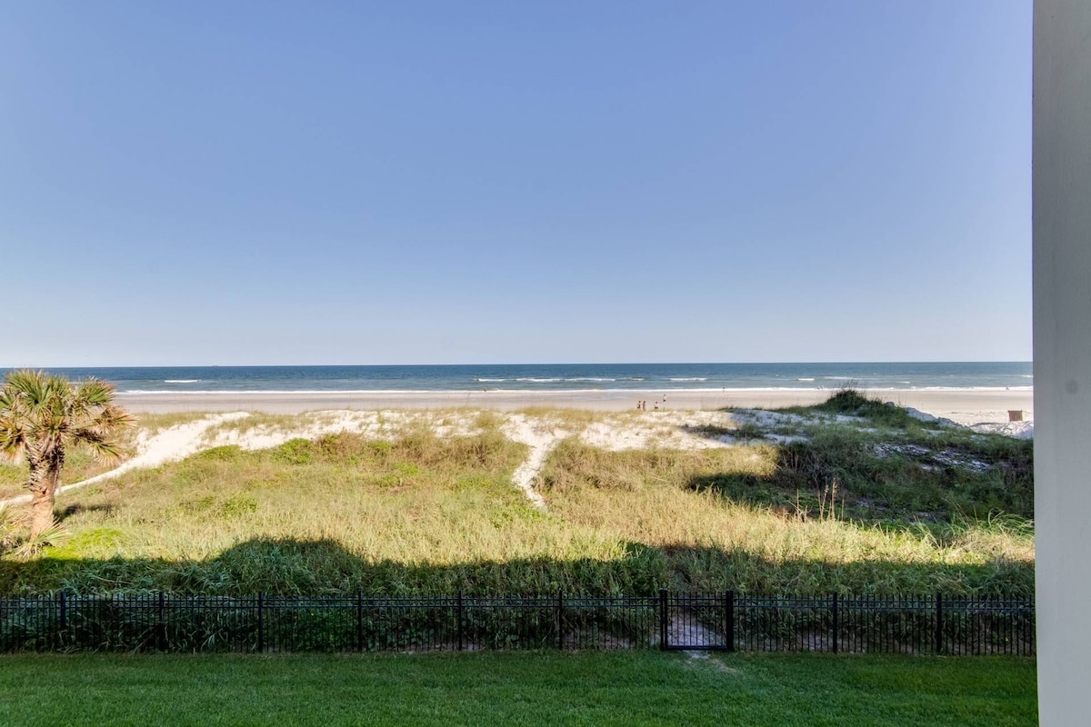 The view captures a serene expanse of sandy beach, framed by lush green grasses. Gentle waves of the Atlantic Ocean can be seen in the distance, while a clear blue sky stretches overhead. A black fence marks the boundary of the outdoor space.