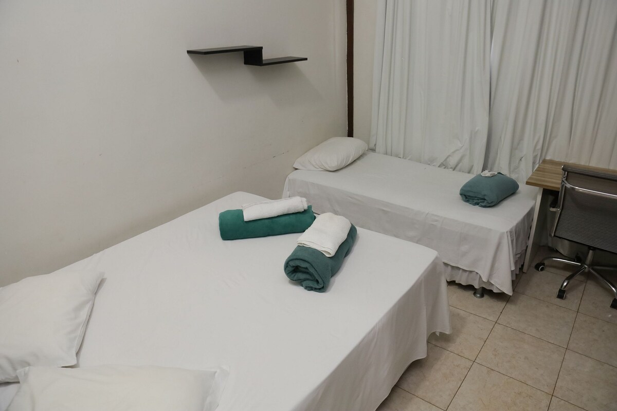 The sleeping area contains two beds dressed in white linens, with light blue towels neatly arranged. A simple black shelf is mounted on the wall, while a workstation with a chair is positioned nearby. Natural light filters through the white curtains.