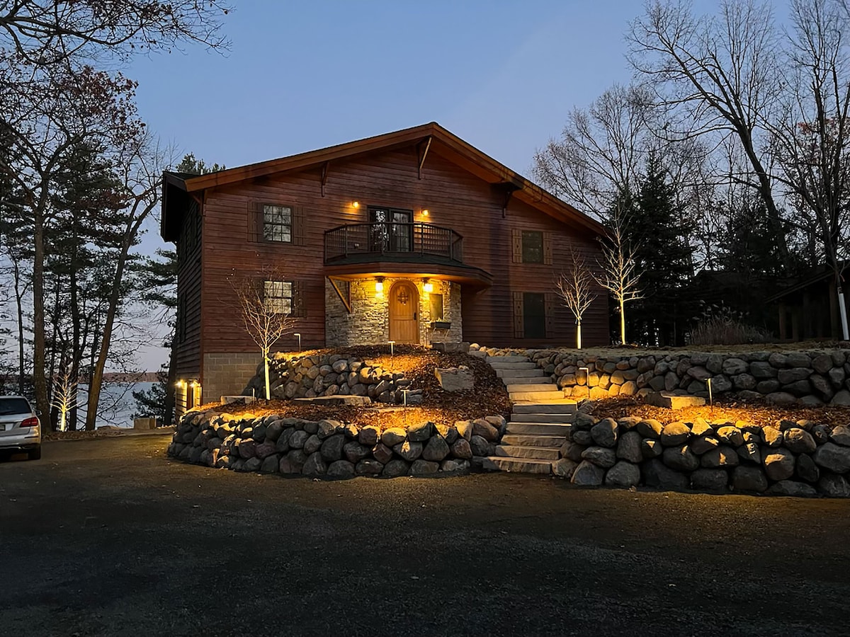 The exterior of the Round House is illuminated by soft outdoor lighting, showcasing its wooden facade and stone accents. A landscaped path leads to the entrance, framed by decorative rocks and trees. The serene lake can be glimpsed in the background.