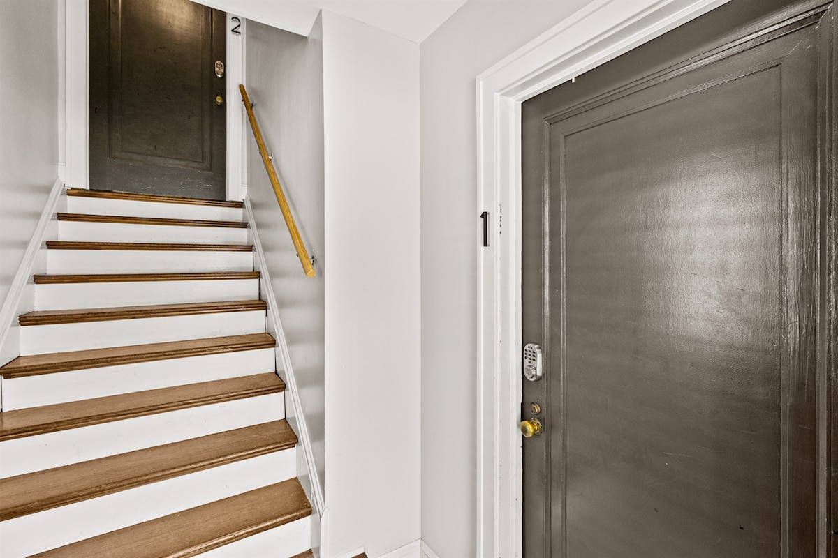A well-lit entryway is depicted, featuring stairs with wooden treads and white risers. A dark front door is visible at the end of the hallway, with a doorbell and keyless entry system installed. The walls are painted a soft gray, creating a clean and modern look.