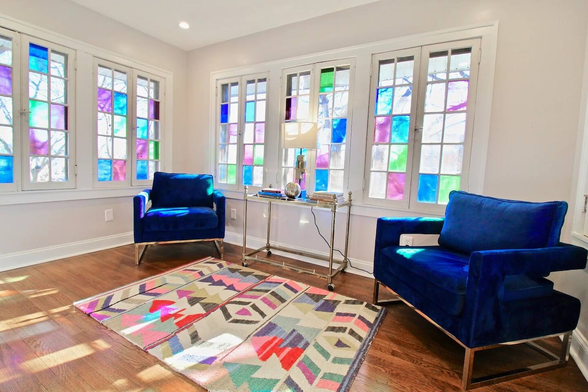 A sunlit seating area is presented, featuring two blue chairs next to a sleek table. Colorful stained glass windows illuminate the room with vibrant hues. A patterned area rug adds a touch of warmth, complementing the hardwood flooring.