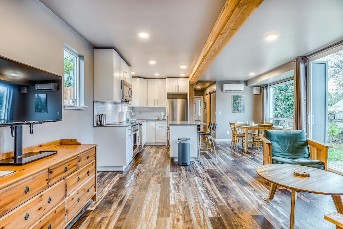 An open living area features modern cabinetry and stainless steel appliances in the kitchen. A wooden dining table is positioned near large glass doors, providing access to outdoor space. The room's warm wood accents are complemented by light streaming in through multiple windows.
