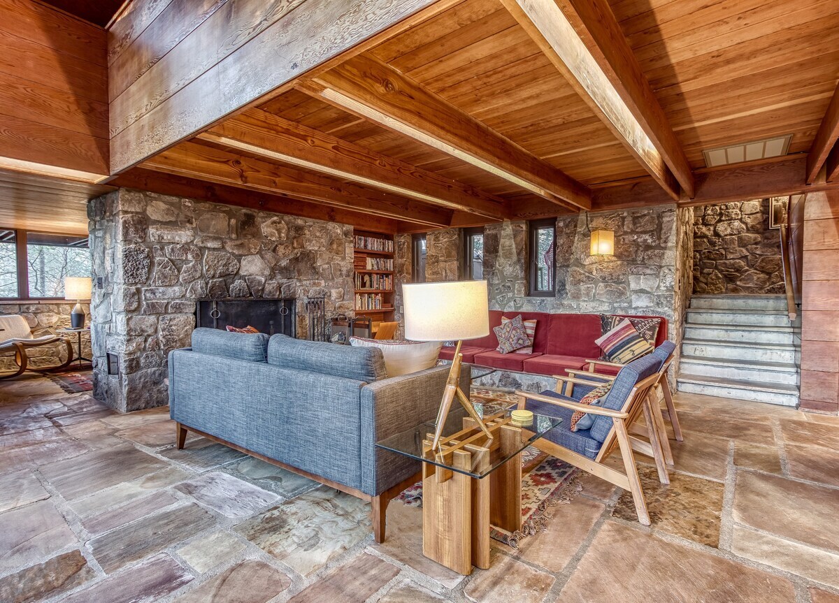 A spacious living room features a blend of stone and wood design elements. Handmade wooden furniture, including a red couch and blue upholstered sofa, surround a glass table. A large stone fireplace serves as the focal point, with warm lighting enhancing the cozy ambiance.
