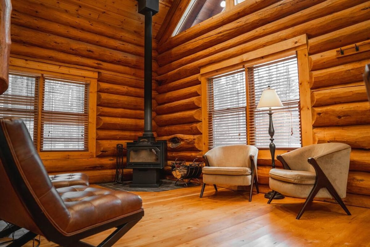 The inviting living area features wooden log walls and a vaulted ceiling. Two armchairs are positioned thoughtfully near a cozy wood stove. Natural light filters through the windows adorned with horizontal blinds, enhancing the warmth of the space.