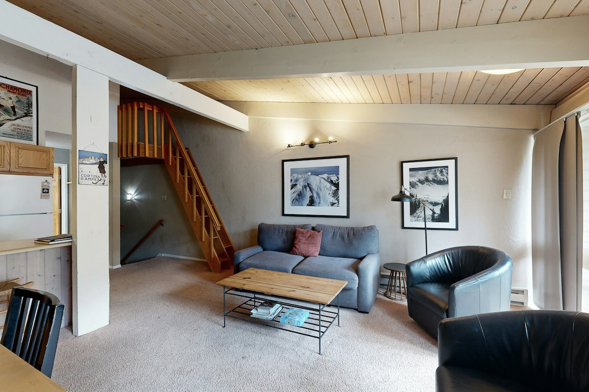 The living area features a spacious sofa, two armchairs, and a wooden coffee table. A staircase leads to an upper level, while large windows allow natural light to brighten the space. Artwork depicting mountain landscapes decorates the walls.