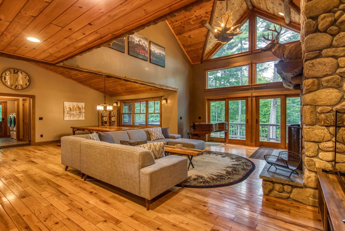 A spacious living room features a sectional sofa positioned on a decorative area rug. Large windows provide views of the wooded surroundings. A stone fireplace is present, complemented by wooden beams and vaulted ceilings, creating a warm and inviting atmosphere.
