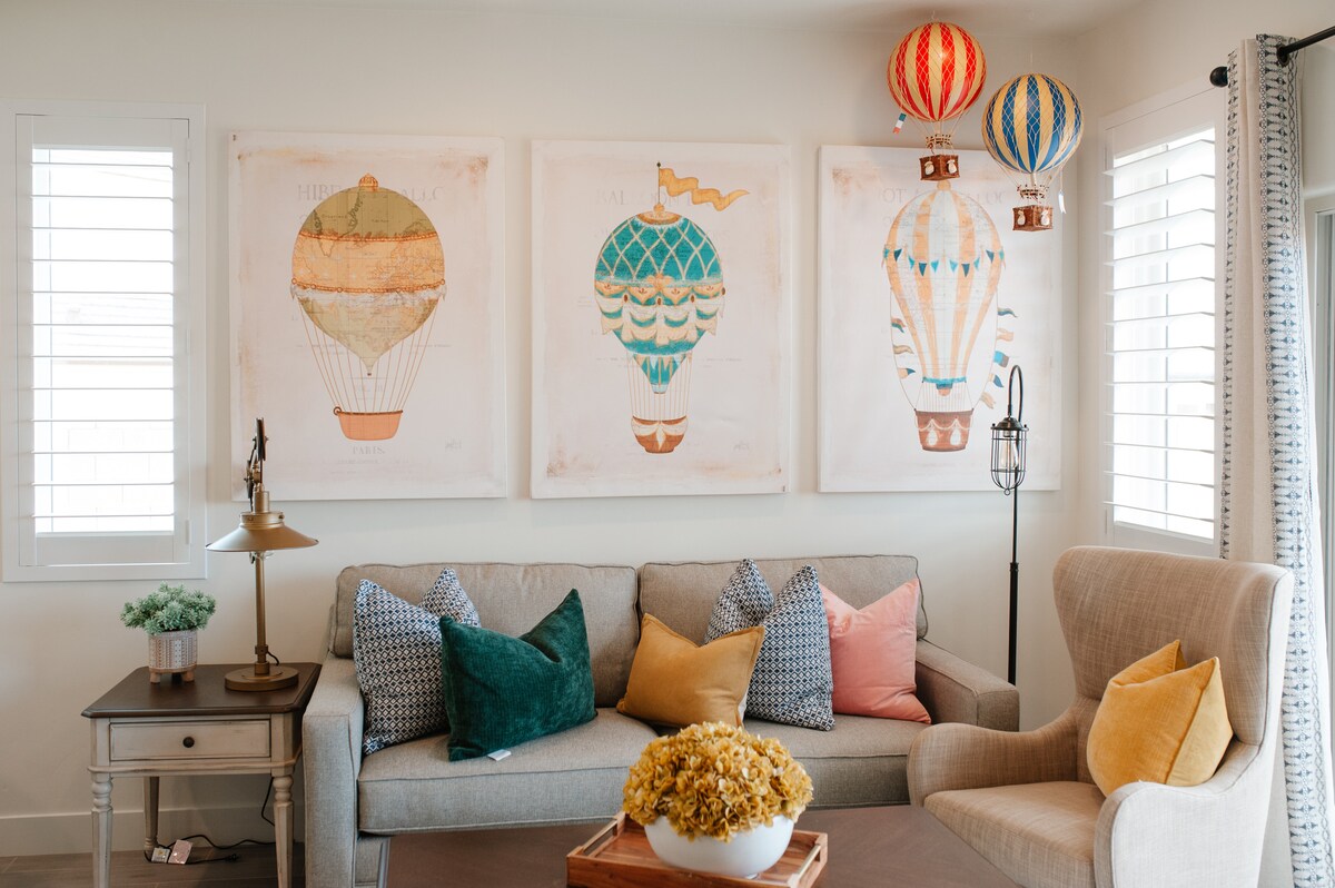A comfortable living area features a light grey sofa adorned with colorful throw pillows. Three framed hot air balloon artworks hang on the wall, complemented by hanging decorative balloons. A wooden coffee table holds a vase of dried flowers, adding a touch of charm.