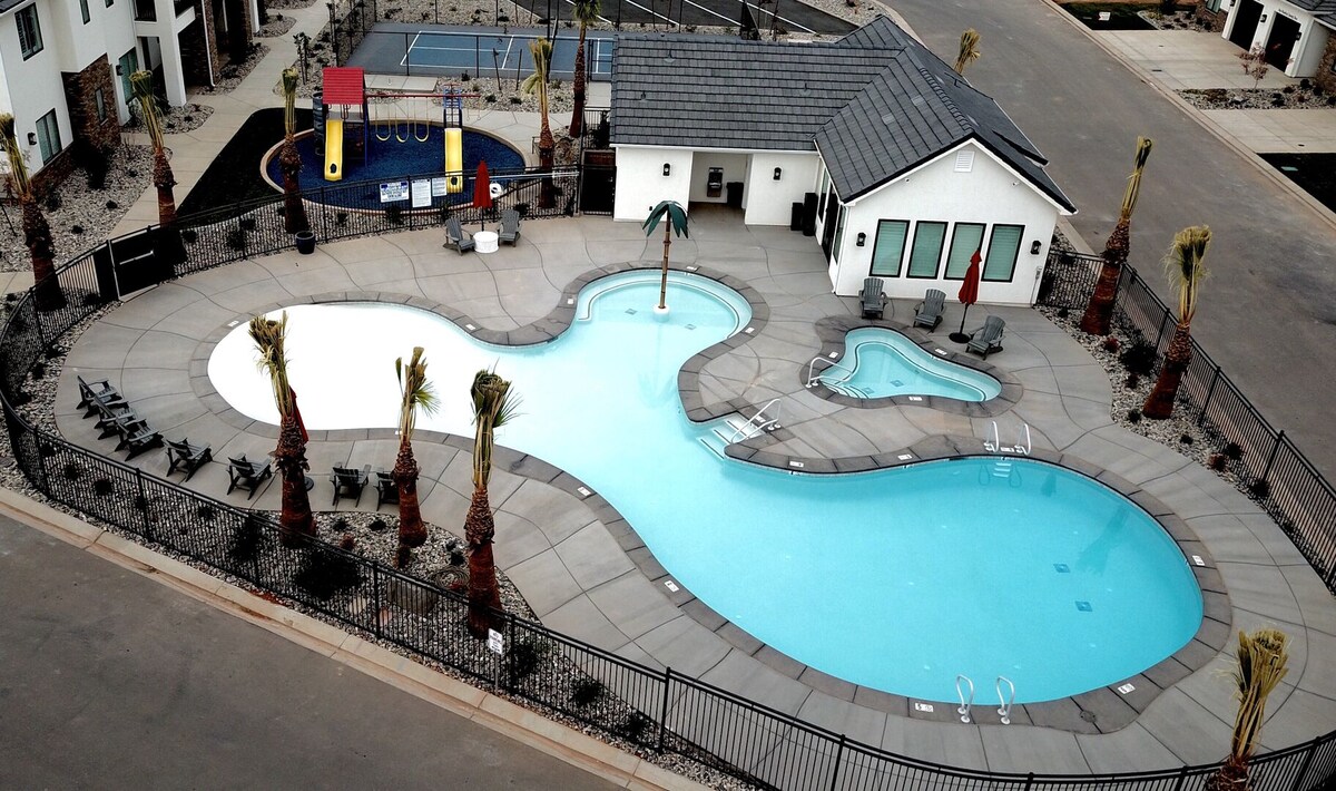 An aerial view of a resort-style pool area is presented, featuring a curved swimming pool, a separate hot tub, and a children's play area with colorful playground equipment. Palm trees border the space, while lounge chairs are positioned for relaxation around the pool's edge.