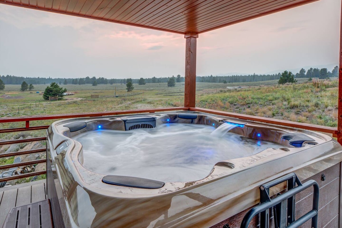 A hot tub positioned on a covered deck offers a serene view of the surrounding landscape. Soft blue lights illuminate the water, while gentle jets create a relaxing ambiance. The open area beyond features grassy fields and distant trees, enhancing the connection to nature.