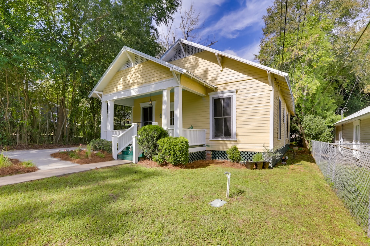 A charming cottage exterior is presented with a light yellow facade, trimmed in white. The covered front porch, flanked by greenery, invites guests. A gravel driveway leads to the side, while a manicured lawn offers space for outdoor activities.