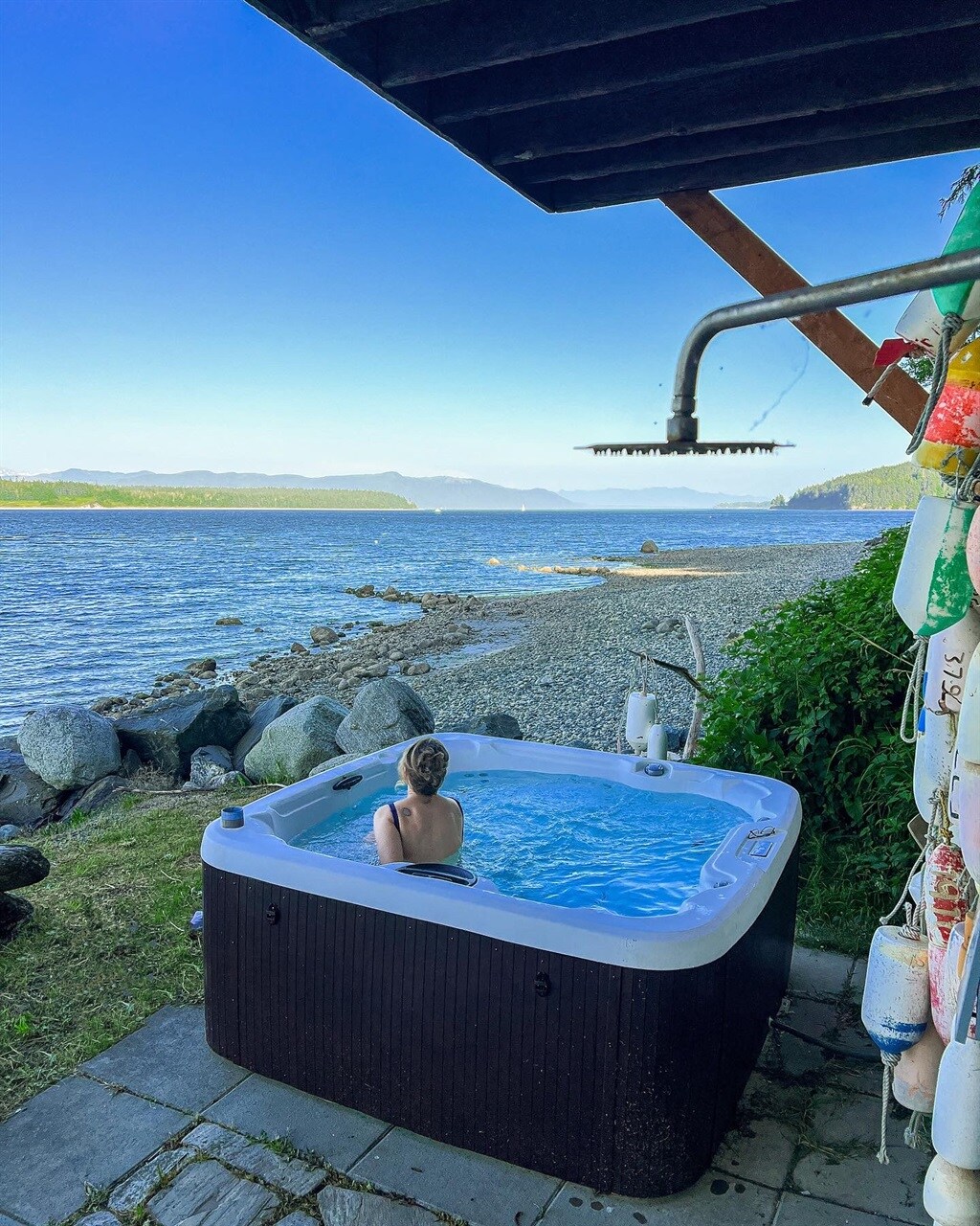 A hot tub is positioned on a stone patio, overlooking the serene beach and water. The surrounding landscape features rocky shorelines and grassy areas, with mountains visible in the distance. A shower head is mounted nearby, allowing for rinsing off after a swim.