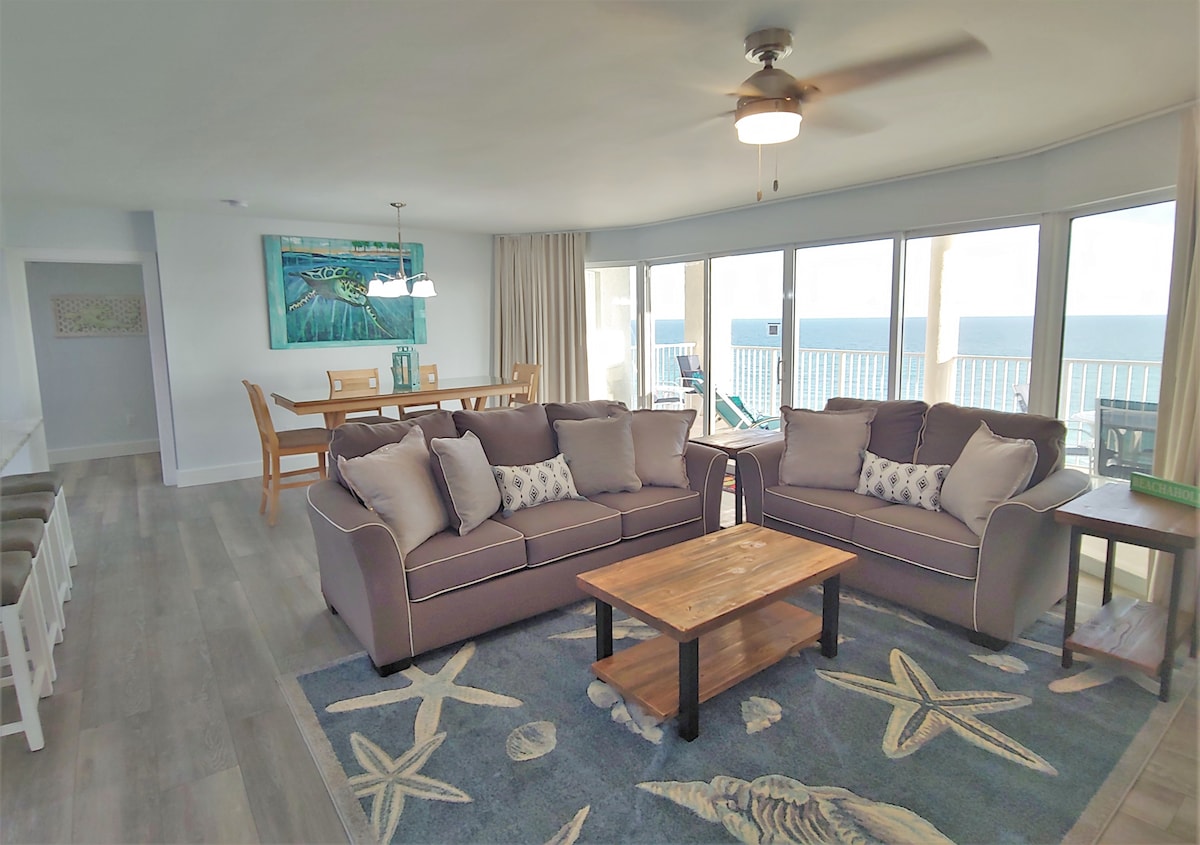 A spacious living area features two comfortable sofas adorned with decorative pillows, complemented by a wooden coffee table. Large floor-to-ceiling windows provide panoramic views of the ocean, while natural light enhances the airy atmosphere. A dining table for six is visible in the background.