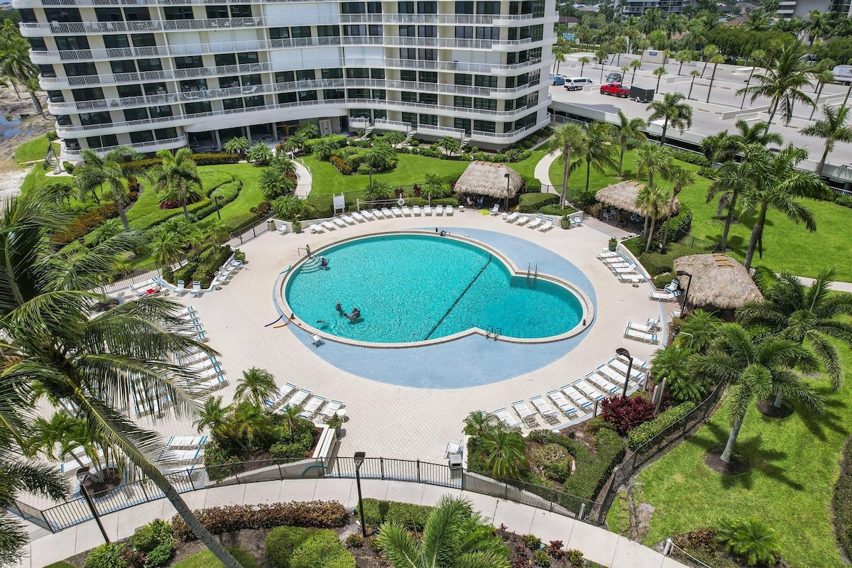 A large heated swimming pool is surrounded by well-maintained tropical landscaping, lounge chairs, and thatched-roof cabanas. The pool's circular shape and inviting turquoise water provide a serene spot for relaxation. A view of the condo building and nearby palm trees can be seen in the background.