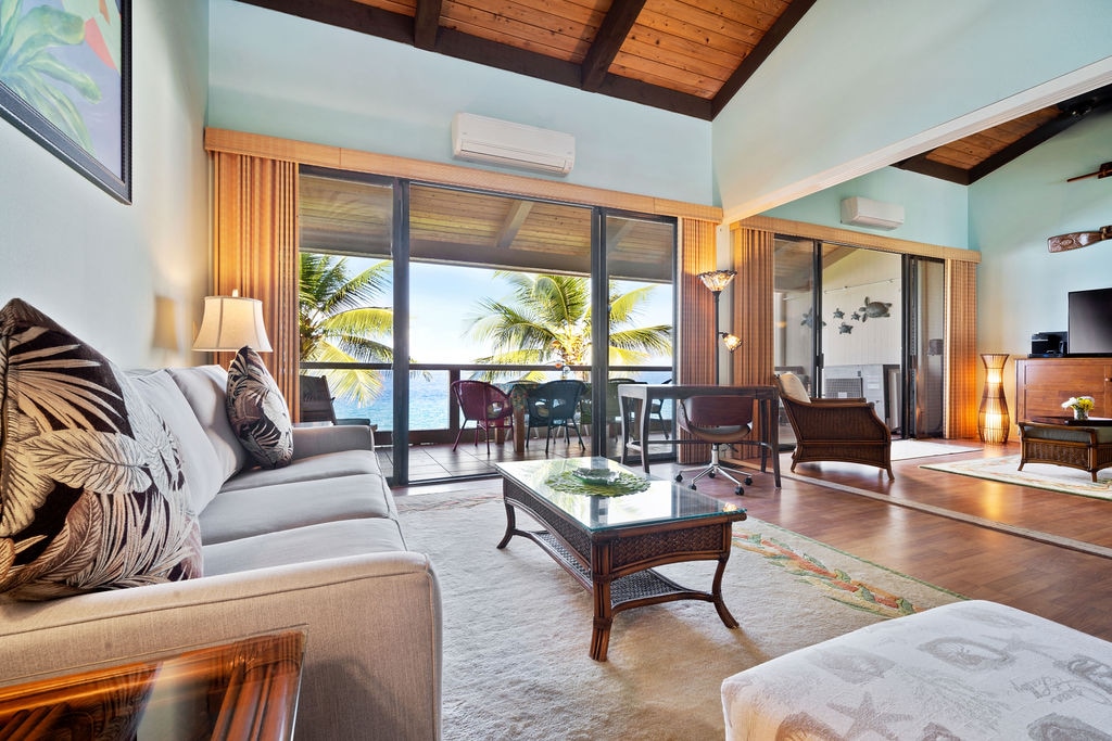 The spacious living area showcases a comfortable sofa and a glass coffee table, with large sliding doors revealing ocean views and palm trees. Soft lighting complements the wooden ceiling, while a dining table is visible on the lanai, enhancing the open and airy feel.
