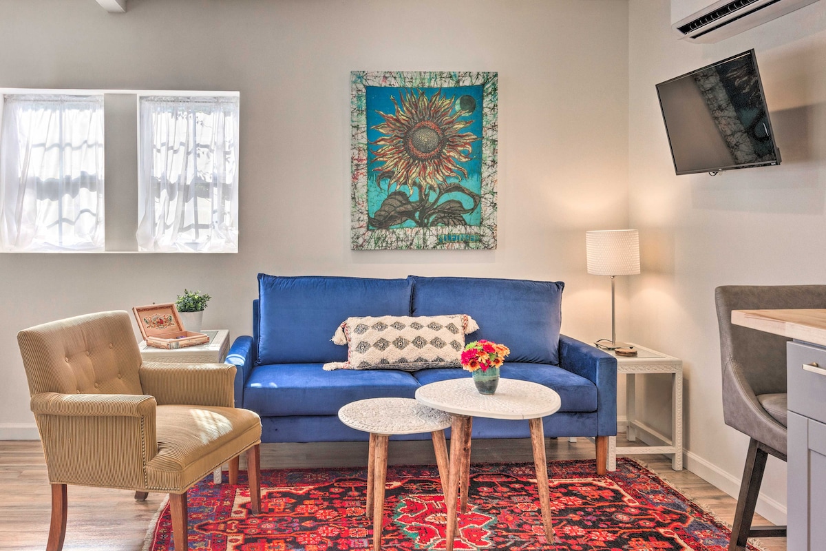 A modern living area features a bright blue sofa with decorative pillows and a coffee table adorned with fresh flowers. Two chairs are situated near the sofa, while an art piece of a sunflower hangs on the wall. Natural light enters from two windows covered with sheer curtains.