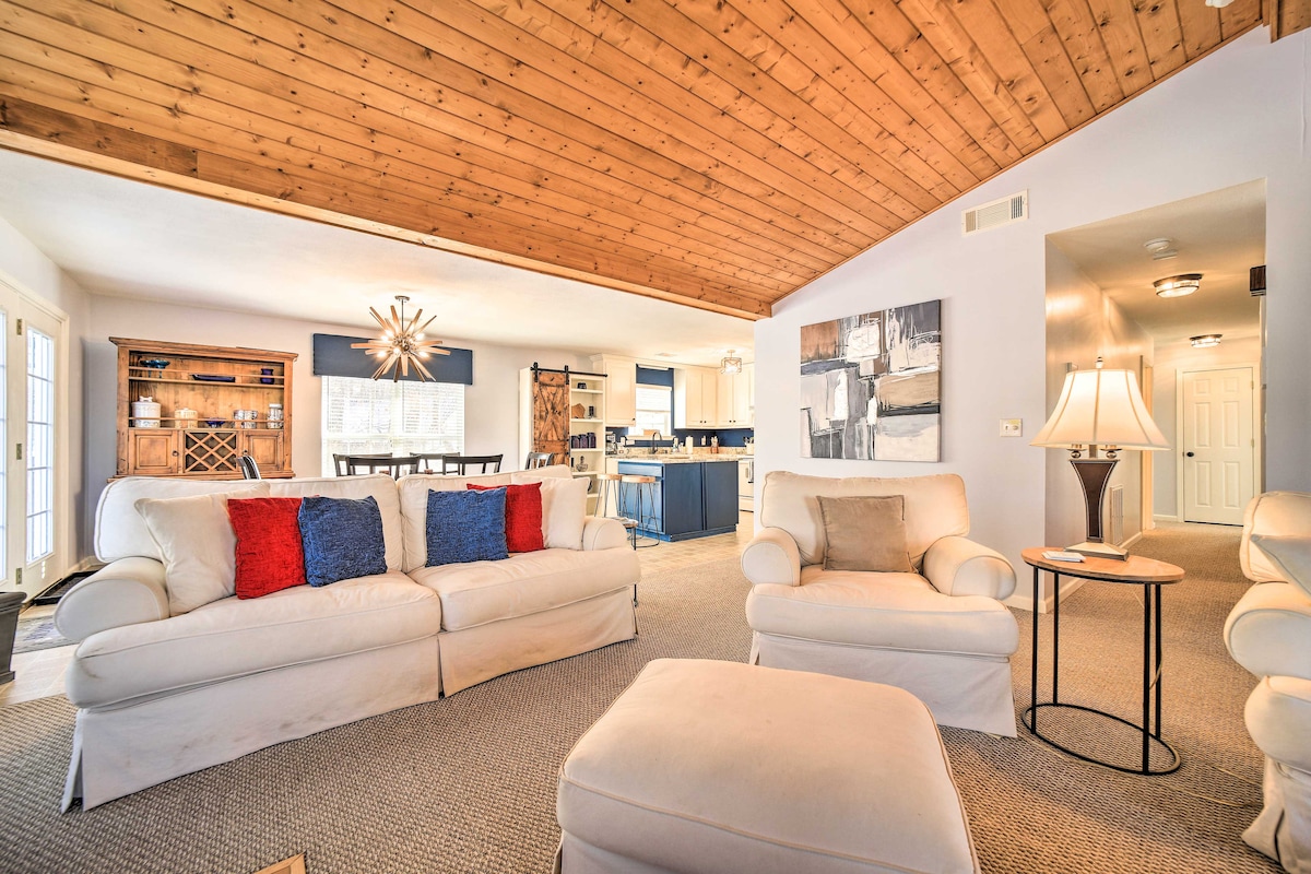 A spacious living area features a vaulted wood-paneled ceiling and cozy seating with light-colored sofas accented by red and blue pillows. A central ottoman is present, and natural light filters through glass doors leading to outdoor space. A dining area is visible in the background.