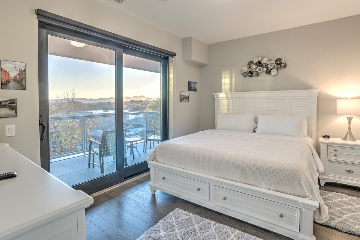 A spacious bedroom features a large bed framed by contemporary bedside tables. Floor-to-ceiling glass doors lead to a private balcony with views of the surrounding landscape. Soft lighting highlights a decorative wall piece above the bed, while a neutral color palette enhances the serene environment.