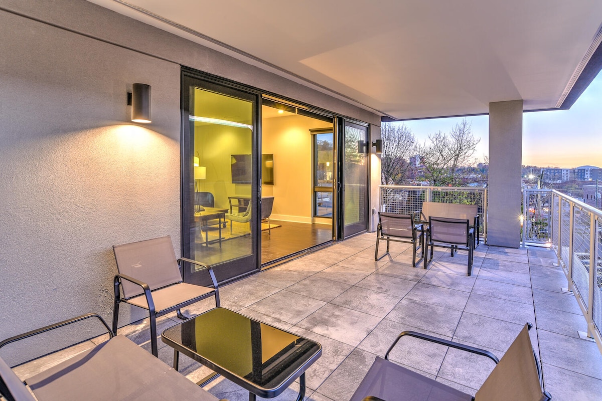 A spacious private balcony is furnished with multiple seating options, including chairs and a table. Large sliding doors provide access to the interior, while soft lighting enhances the serene evening atmosphere. Expansive views of the surrounding area are visible beyond the railing.