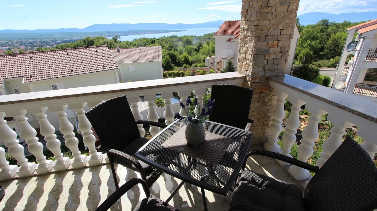 A balcony space features a round table surrounded by four woven chairs, with a vase of lavender at the center. Scenic views of the sea and distant hills are visible in the background, framed by the decorative stone columns of the balcony.