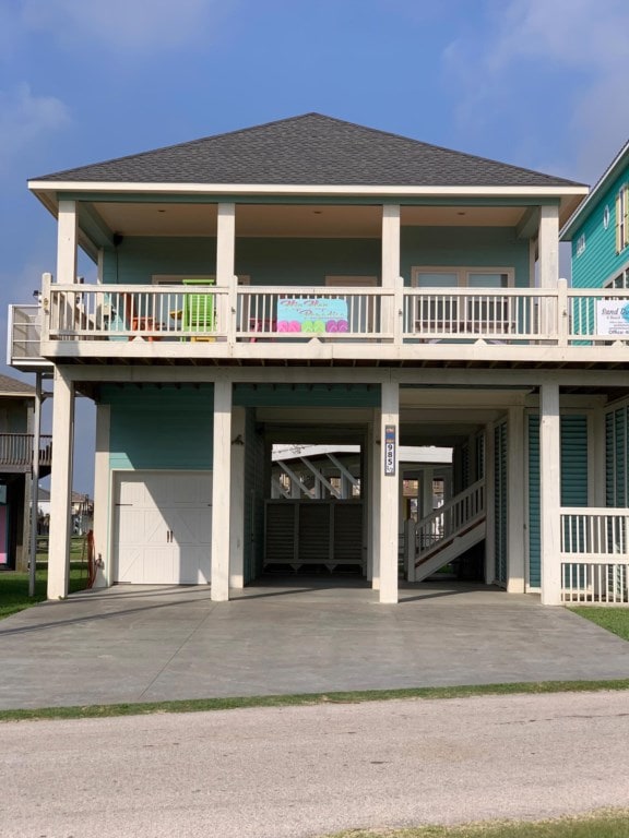 A two-story beach home is showcased with a light blue exterior. The large covered balcony is complete with white railings, offering seating options. A spacious ground-level area is visible, with a driveway leading to an enclosed space underneath the house.