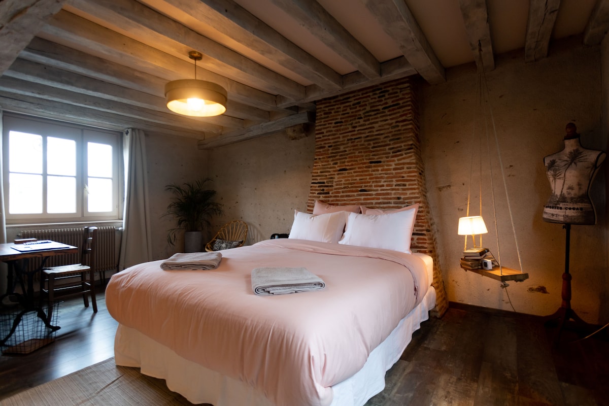 A spacious bedroom features a large pink bedspread atop a comfortable bed, complemented by textured plaster walls and wooden beams overhead. A vintage mannequin stands beside a decorative lamp, with a small table and chair arranged near the window that lets in natural light.