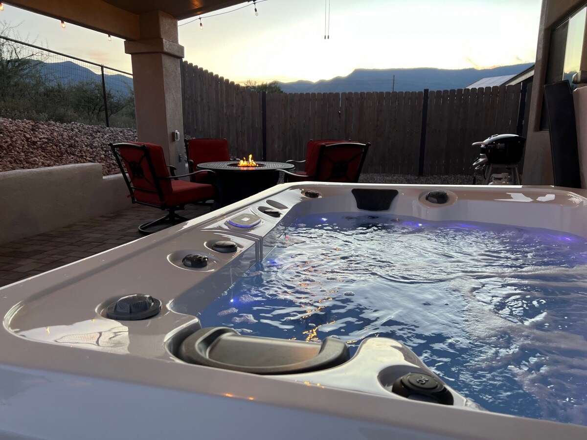 A private outdoor hot tub is shown with gently bubbling water, surrounded by a spacious patio area. A firepit, two comfortable red chairs, and a small table are visible in the background, with a wooden fence and mountains visible beyond.