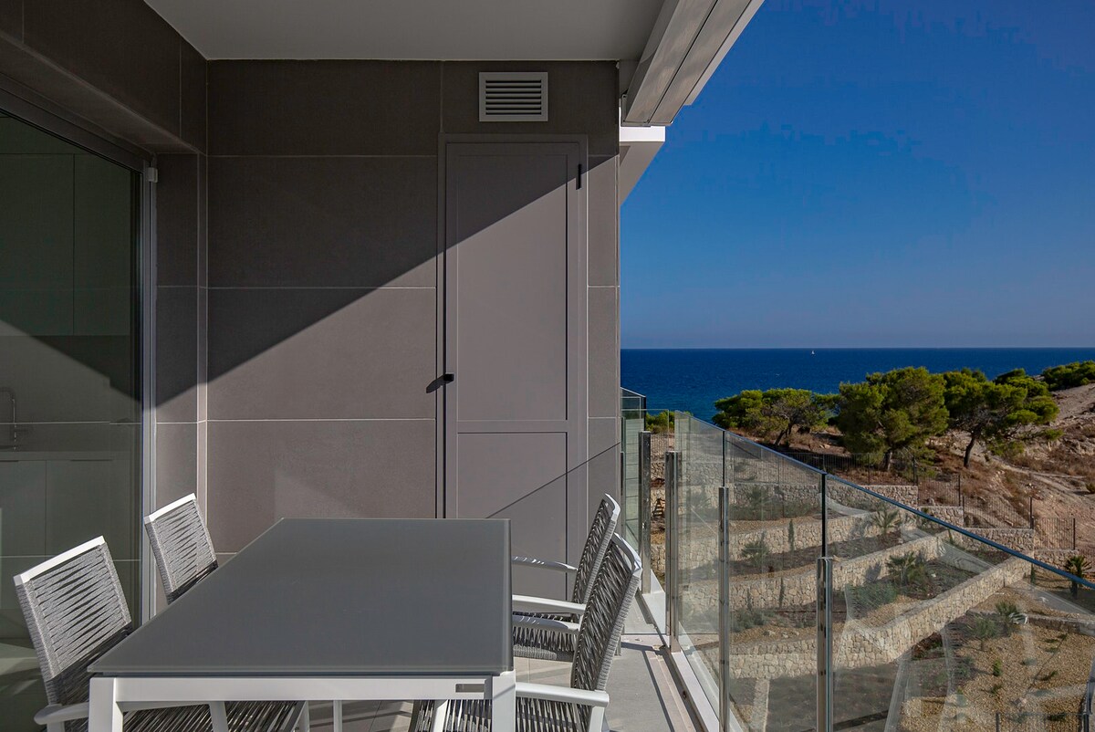 A spacious terrace features a modern dining table with several matching chairs. A glass railing provides unobstructed views of the blue sea and surrounding landscape, with a clear blue sky above. The contemporary architecture complements the serene outdoor setting.