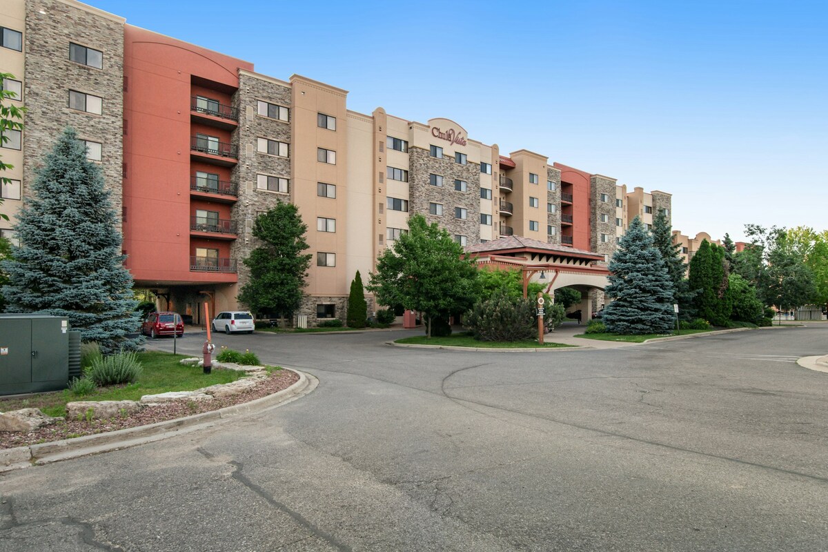The exterior of the Chula Vista Resort Condo is presented, featuring a multi-story building with a mix of stone and stucco finishes. Lush greenery surrounds the entrance, and vehicles are parked along the driveway, with trees adding to the natural ambiance of the setting.