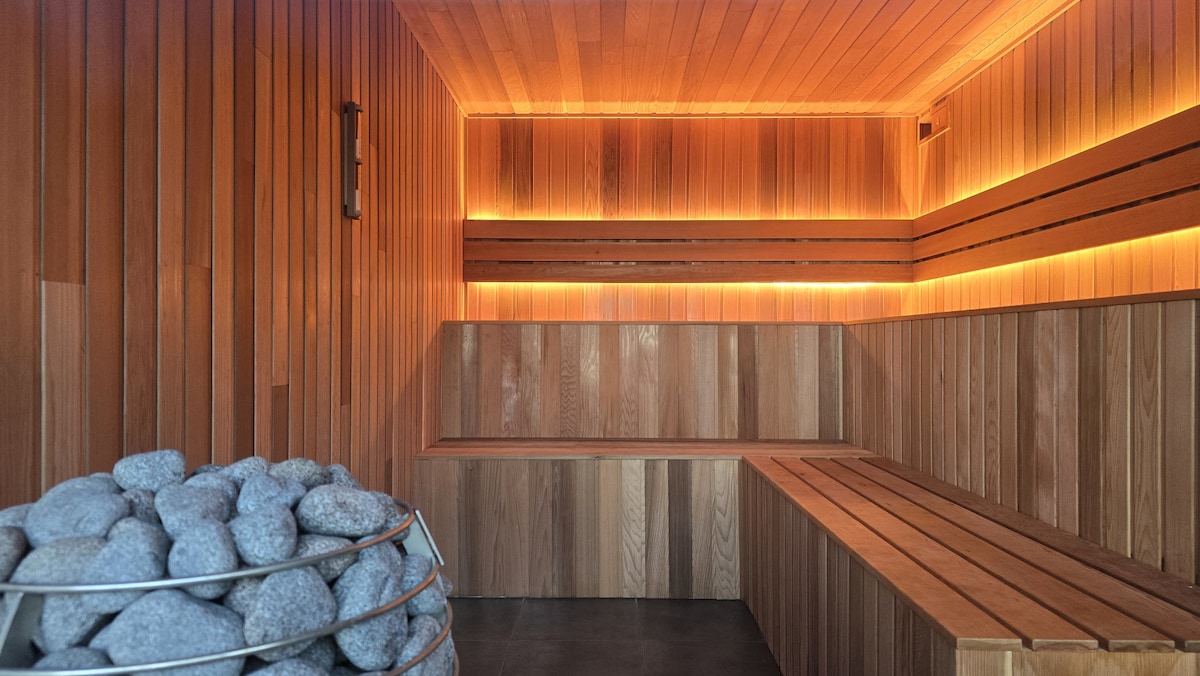 A wooden sauna interior is illuminated by soft, warm lighting. Smooth wooden benches line the walls, while a stack of rounded stones is placed in a metal basket. The wood paneling creates a calming atmosphere, enhancing the spa-like experience.