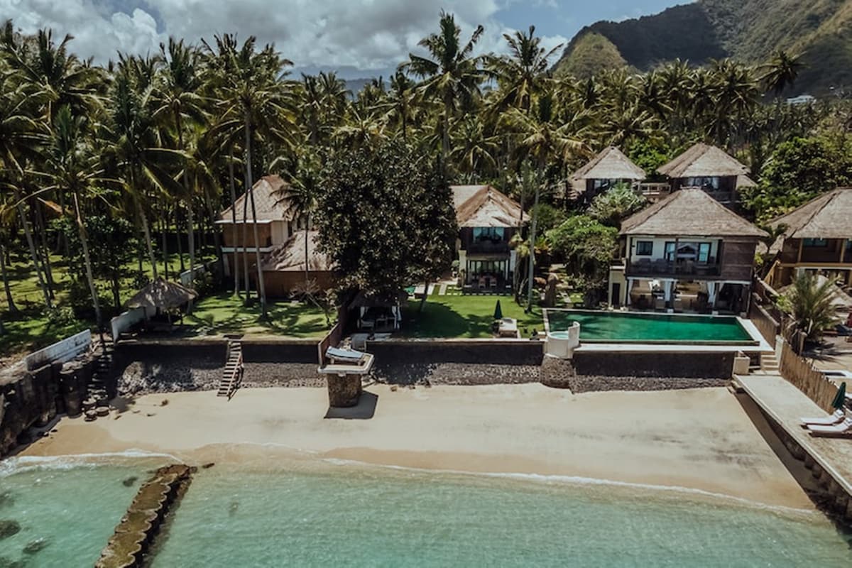 An aerial view of a tropical resort along the shoreline showcases several villas surrounded by palm trees. A clear turquoise pool is visible near the ocean, with a sandy beach in the foreground. The lush green landscape offers a serene atmosphere.