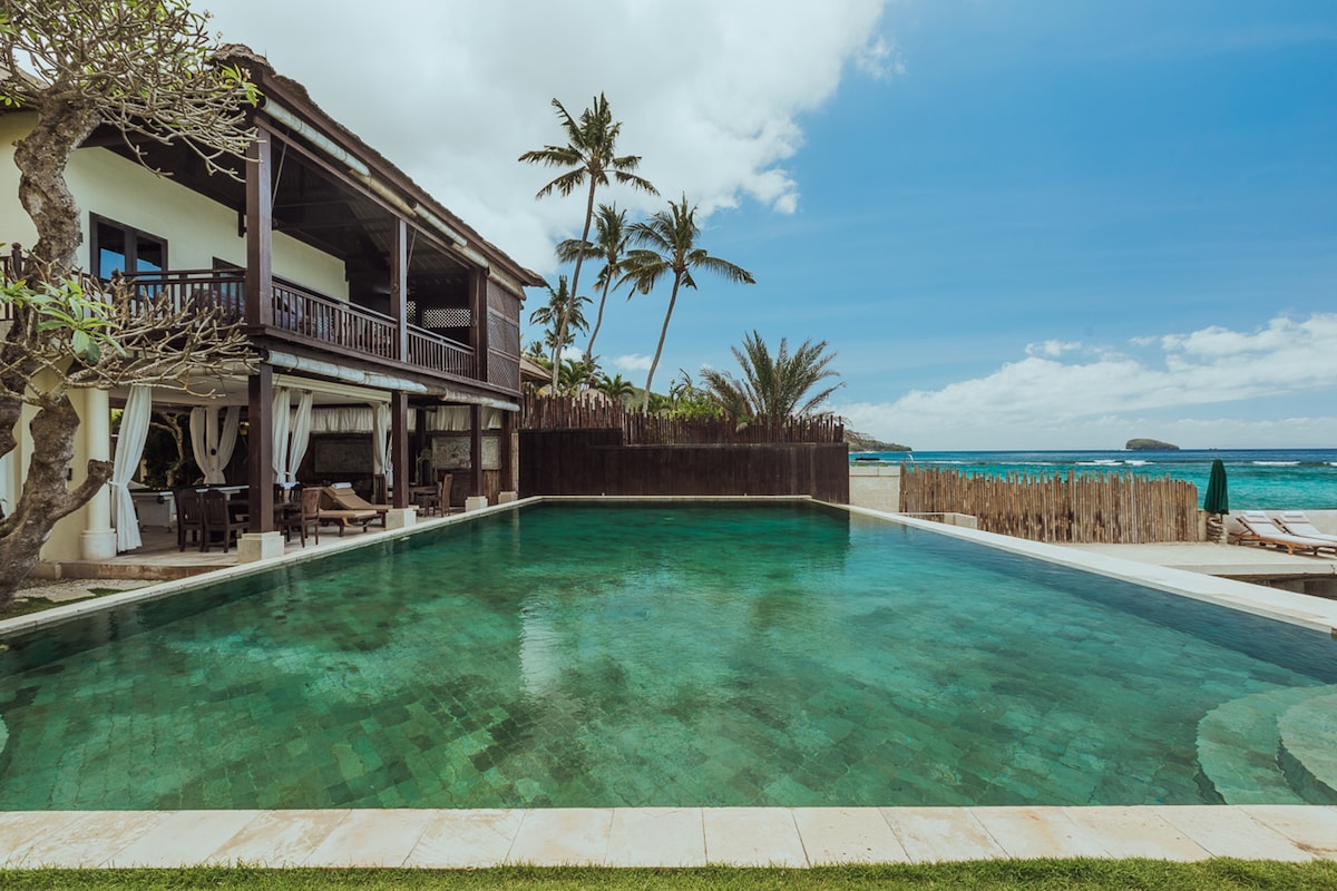 A spacious swimming pool is set against the backdrop of a two-story villa, with expansive wooden balconies and tropical palm trees surrounding the area. The clear blue sky enhances the inviting atmosphere of the pool, which reflects the ocean beyond.