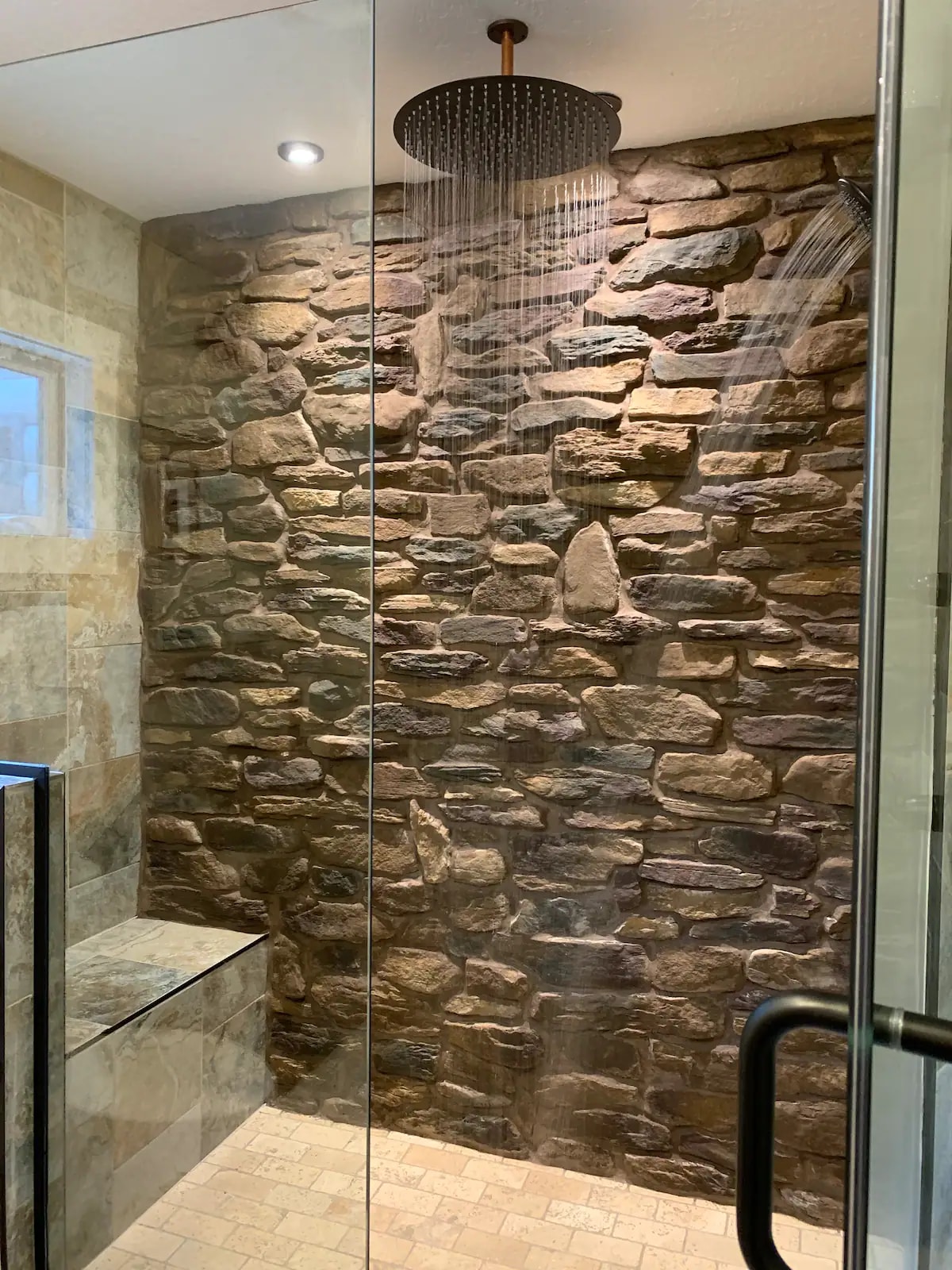 A luxurious rainfall shower is showcased within a stone-walled enclosure. Soft lighting highlights the textured surface of the natural stone, while water cascades down from a sleek overhead showerhead. A built-in bench is visible, providing a space for comfort and convenience.