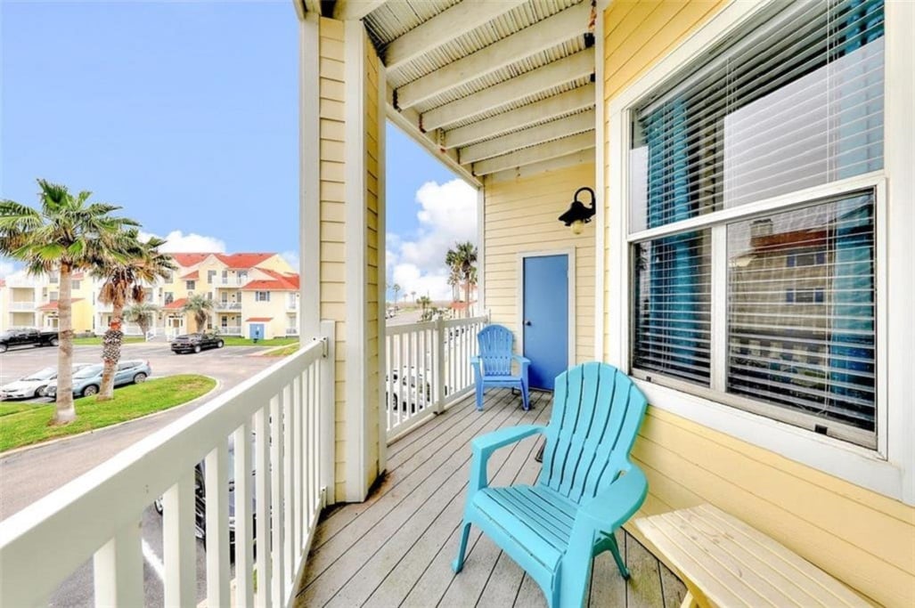 A private balcony features two blue Adirondack chairs, inviting relaxation with a view of the surrounding community. Lush palm trees and vibrant buildings are visible in the background, framed by the clear sky, creating a welcoming outdoor space.