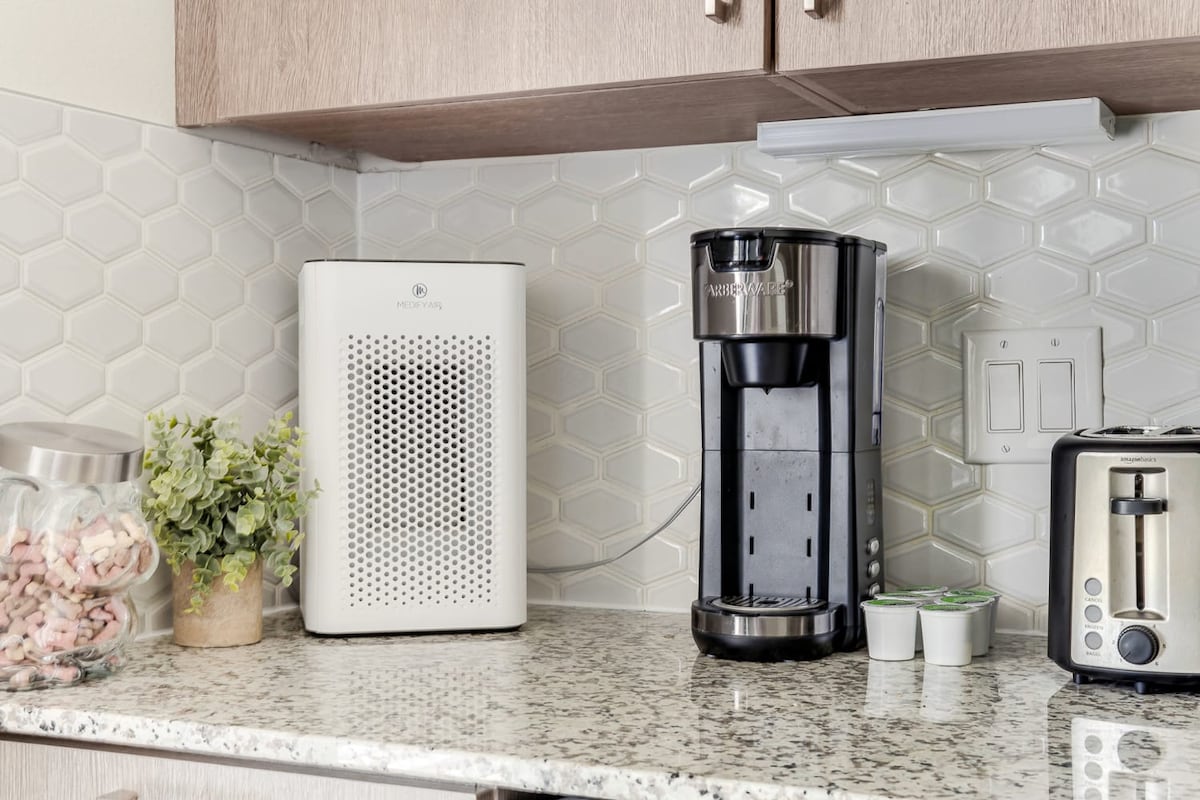 A modern kitchen countertop is displayed, featuring a coffee maker next to a stylish air purifier and a compact toaster. Decorative elements include a glass jar filled with pink marshmallows, and a small potted plant adds a fresh touch. The granite surface complements the hexagonal tiled backsplash.