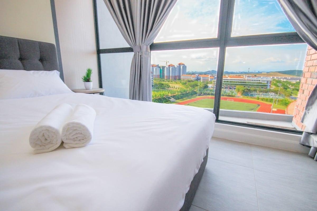 A spacious bed adorned with neatly rolled towels is positioned near large windows. Daylight filters through sheer curtains, illuminating the room. A view of greenery and nearby buildings can be seen outside, creating a serene atmosphere.