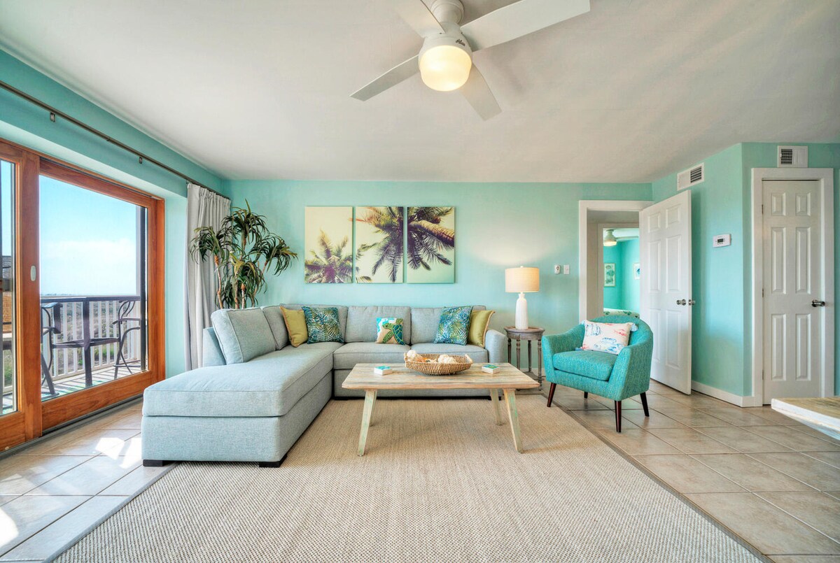The living area is furnished with a spacious light blue sectional sofa and a small accent chair. A wooden coffee table is positioned atop a woven area rug. Large glass doors open to the balcony, allowing natural light to illuminate the soft turquoise walls and tropical artwork.