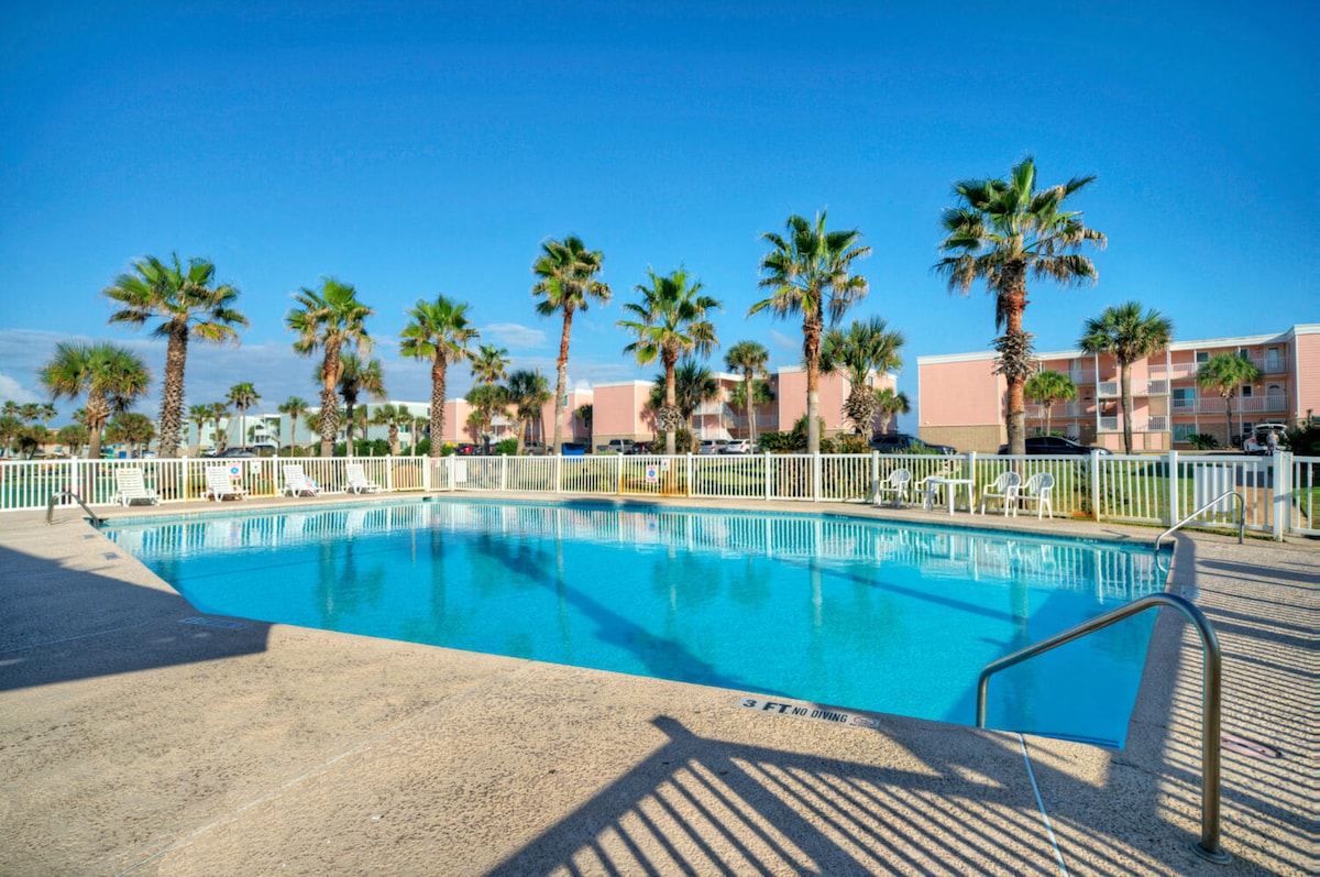 A spacious pool is surrounded by palm trees, creating a serene setting. The clear water reflects the blue sky, and lounge chairs are placed around the perimeter for relaxation. A white safety fence encloses the pool area.